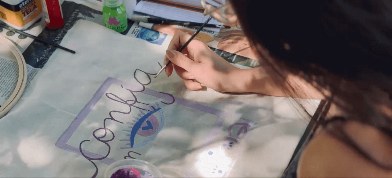 Una persona pintando con pincel una bolsa de tela, en la bolsa pone confía y hay dibujado un ojo